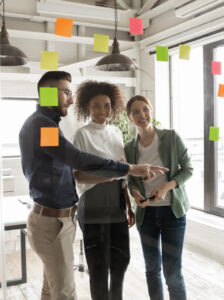 Happy young mixed race diverse business people managers standing near glass wall with pinned colorful sticky notes, managing workflow or organizing project processes together in modern office.